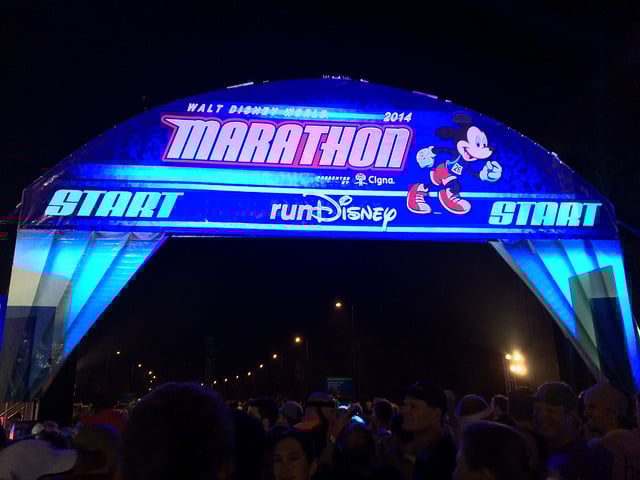 A large crowd is gathered under an illuminated arch at night, signaling the starting line for the Walt Disney World 2014 Marathon. The arch is decorated with an image of Mickey Mouse running and features both the Cigna and runDisney logos.