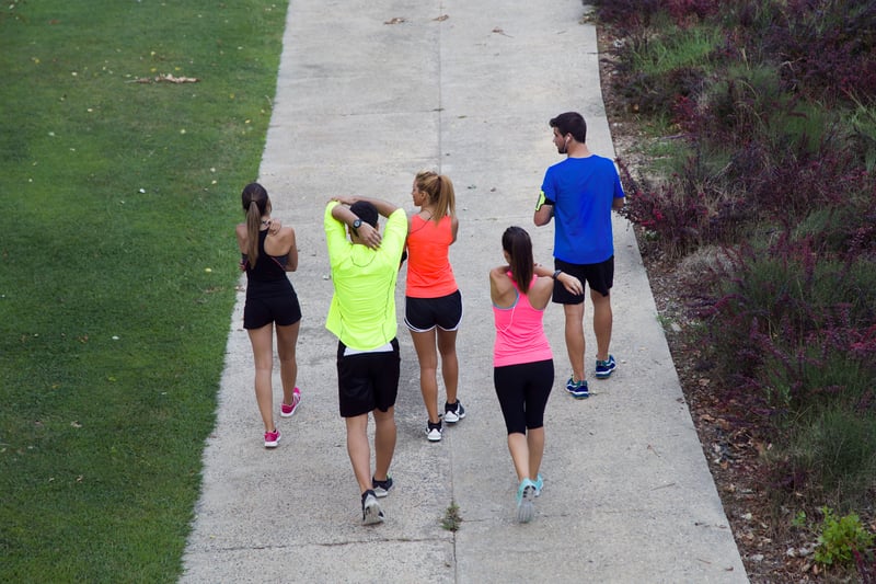 Five people wearing athletic clothing are walking on a paved path outdoors. Three women and two men are seen from behind, with some stretching their arms. On the left is a grassy area, and on the right, there are some shrubs and plants.