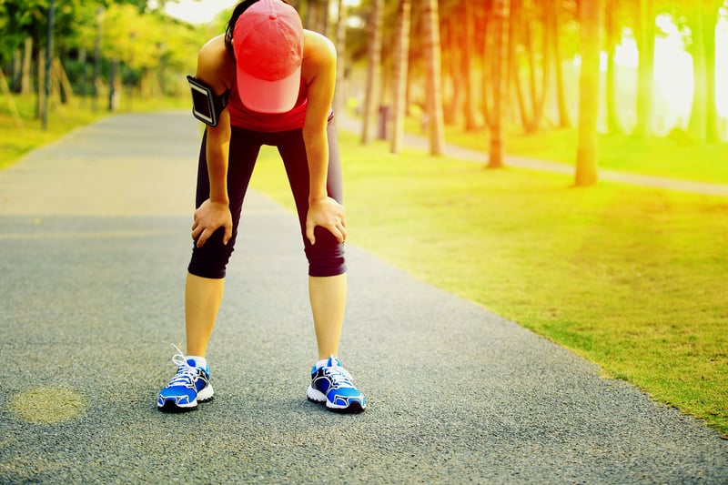 A person wearing a red cap and athletic clothing is bent over with hands on their knees, appearing tired, on a paved path in a park. The sun is shining brightly, creating a warm and glowing background with trees lining the path.