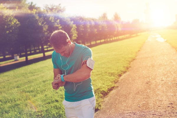 A man in a turquoise shirt and white shorts standing on a path in a park at sunset. He is looking at a smartwatch on his wrist and wearing earphones, with a smartphone in an armband. The sun is casting a warm, golden glow over the scene.