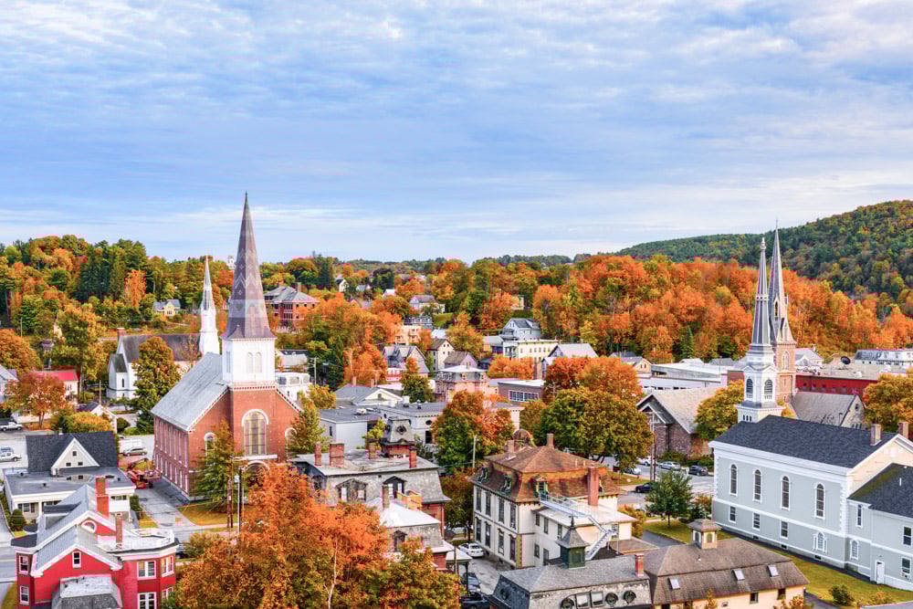 A scenic aerial view of a quaint New England town during fall, showcasing colorful foliage with orange, red, and yellow leaves. The town features charming buildings, churches with tall steeples, and tree-lined streets set against a backdrop of gently rolling hills under a partly cloudy sky—a picturesque setting for half marathons.